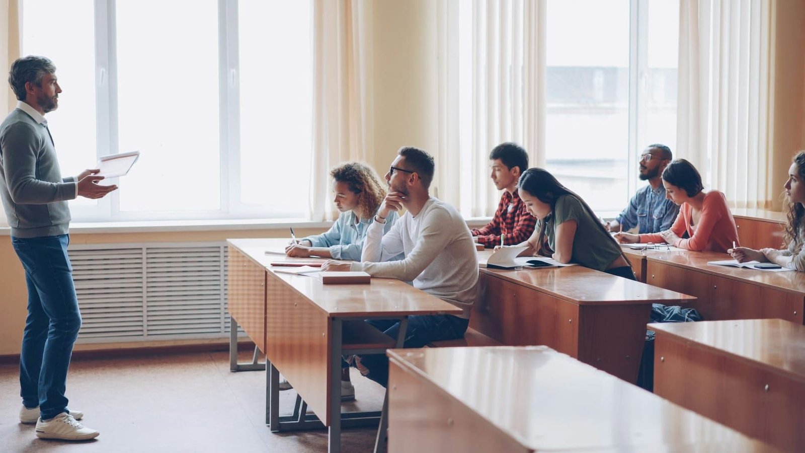 Students seated at long rows of desks in a college classroom while a lecturer stands at the front.