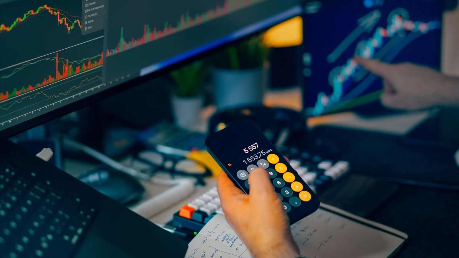 Hands working a desk calculator beside a screen showing financial charts and figures.