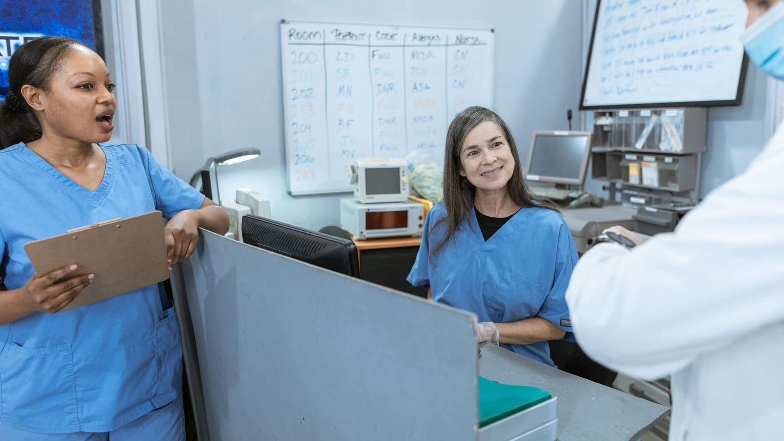 Clinical staff at a hospital nursing station, one holding a patient chart, with a shift whiteboard behind them.