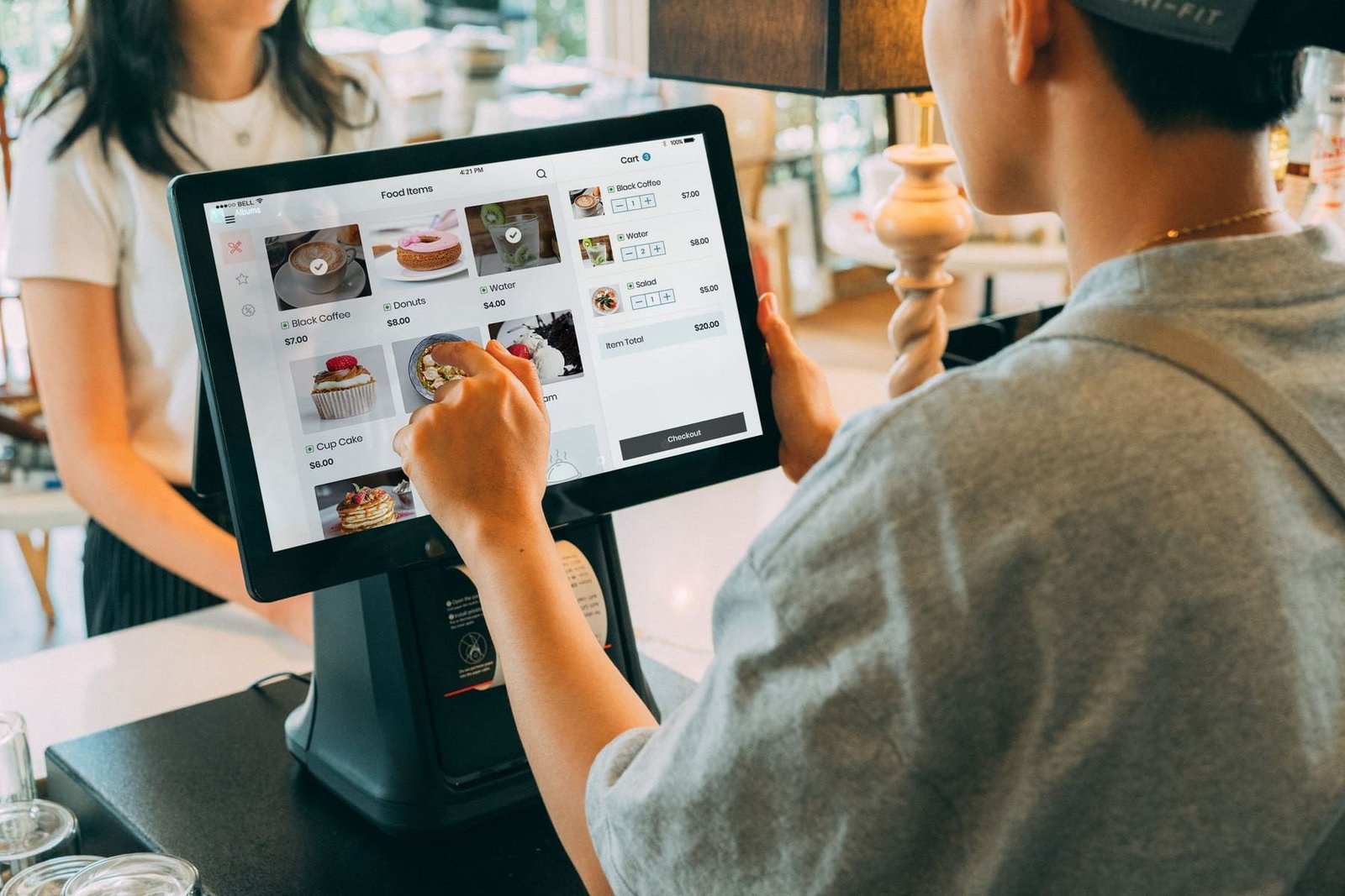 A shop assistant tapping an item on a point-of-sale touchscreen at a counter while a customer waits.