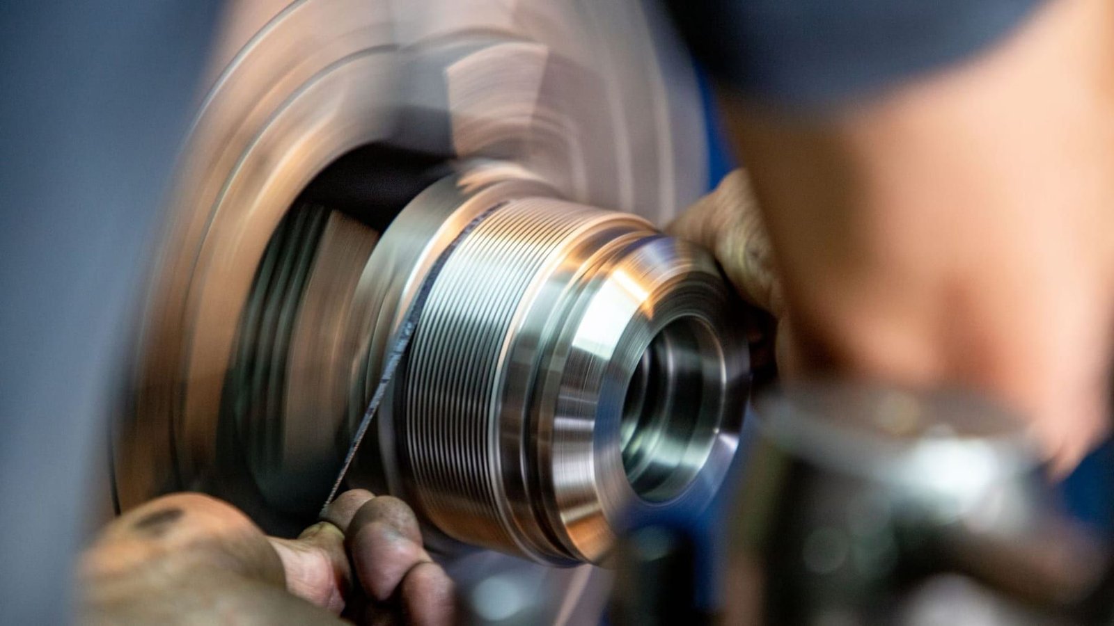 Close-up of a cylindrical steel component spinning in a machine while an operator finishes its surface by hand.