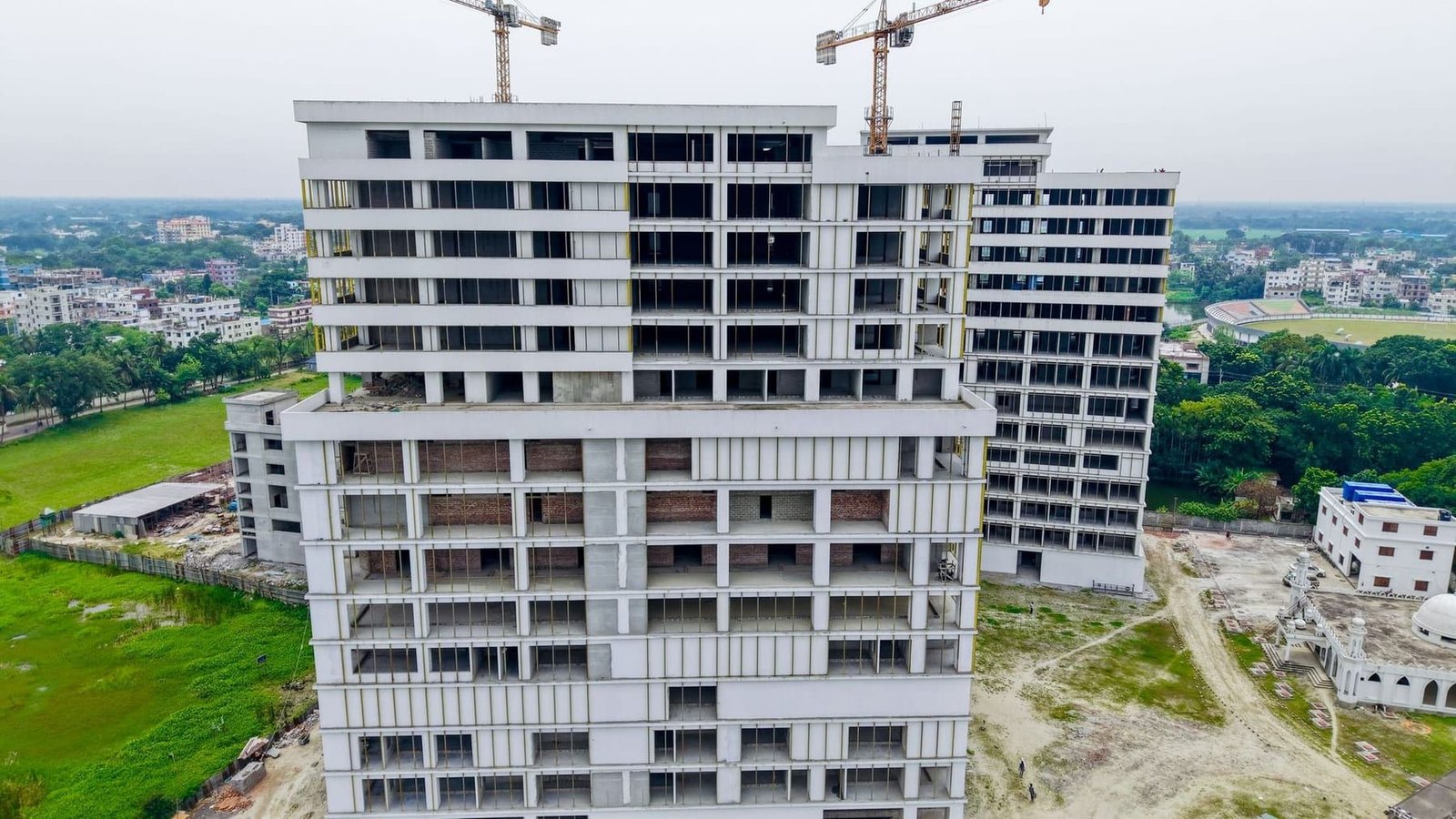 A multi-storey residential block under construction, tower cranes standing over its unfinished upper floors.