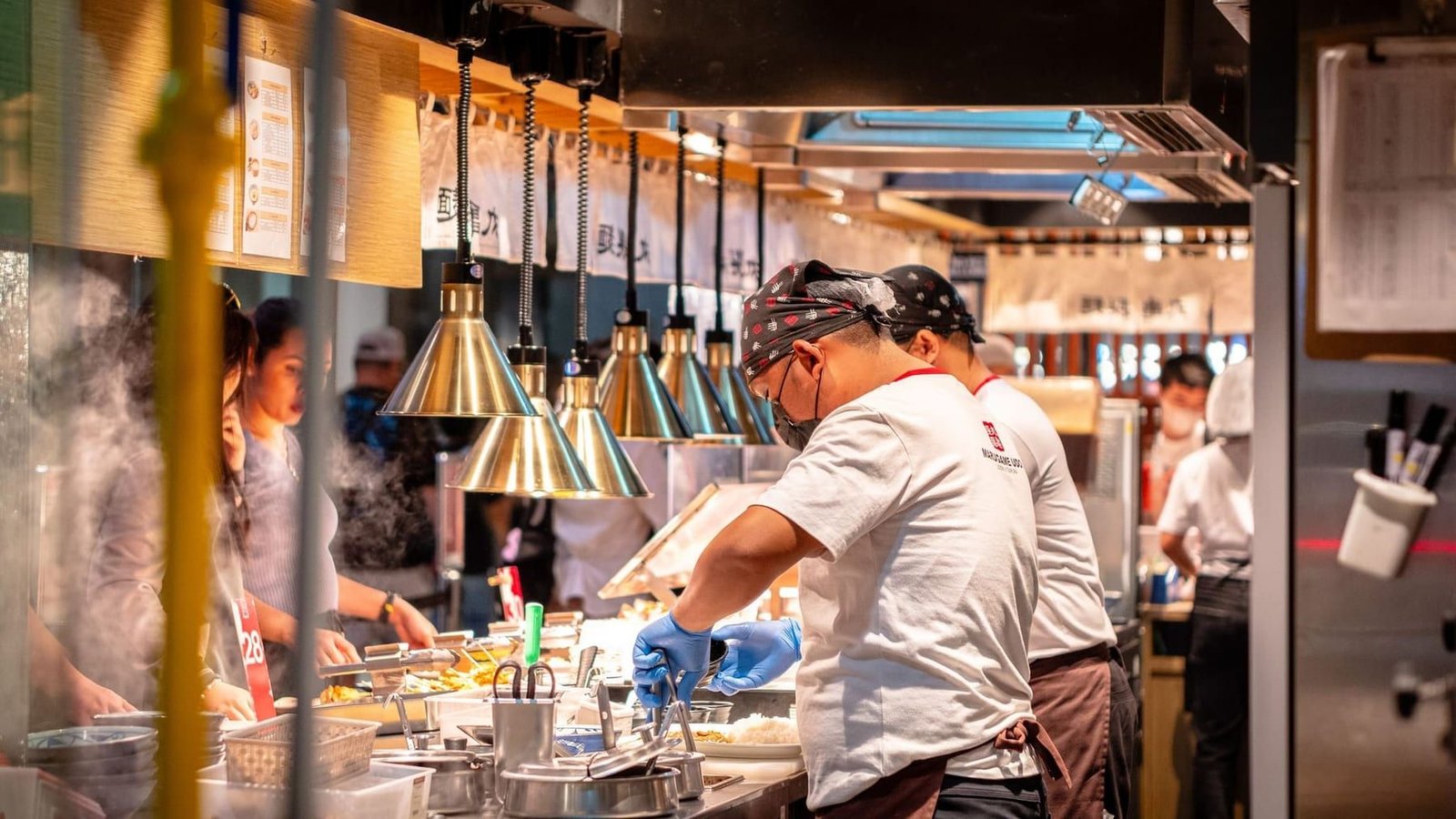 Chefs plating dishes along a restaurant kitchen pass under heat lamps during a busy service.
