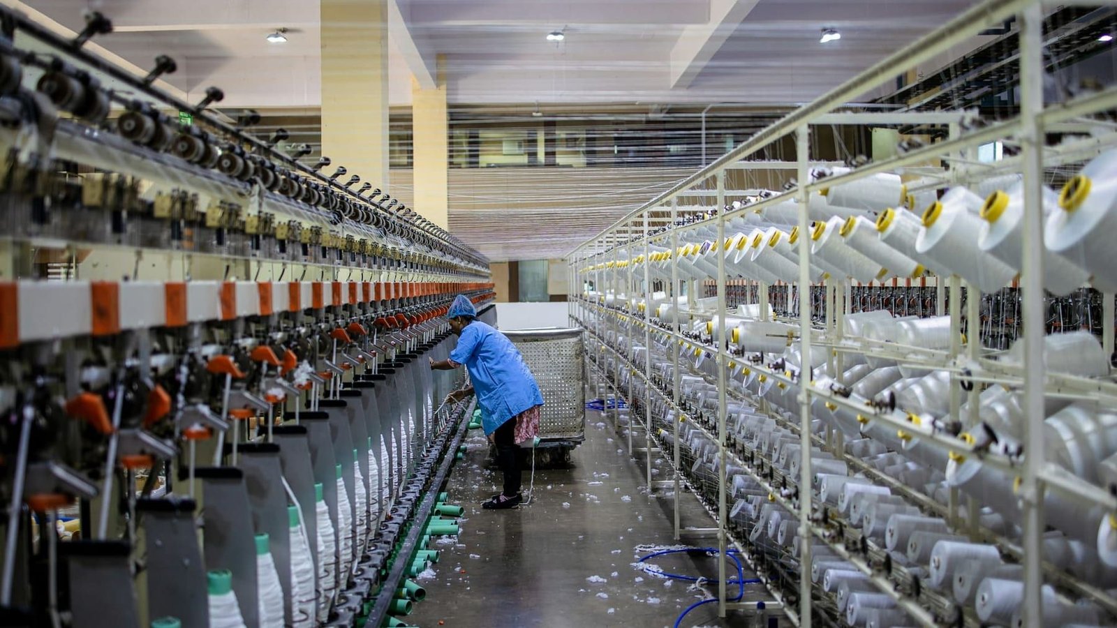 A long row of spinning machines running yarn packages in a textile mill, with a worker tending the line.